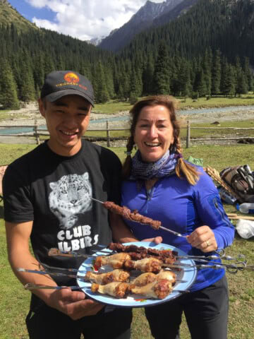 Two people outdoors hold a plate with skewered meat, smiling. The man wears a black hat and T-shirt with a snow leopard. The woman is in a blue jacket with a forest and mountain landscape in the background, reminiscent of the natural beauty found in Kyrgyzstan.