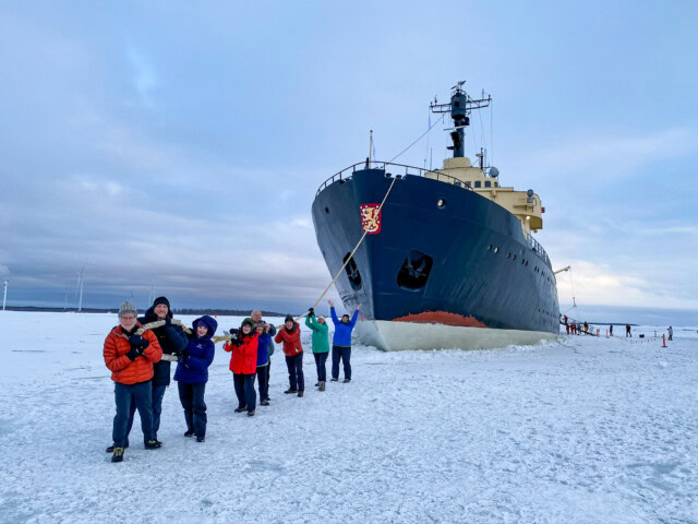 A group of people stand on ice in front of a large, docked ship in Finland, posing for a photo in winter attire. The sky is overcast and the ship's bow is prominently visible.