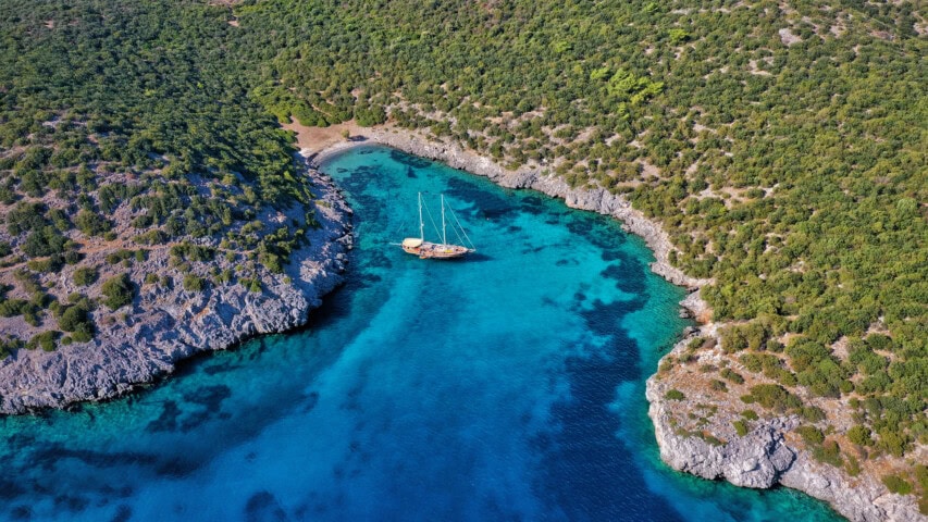 Aerial view of a sailboat anchored in a secluded, clear blue water bay surrounded by dense green vegetation and rocky cliffs in Turkey.