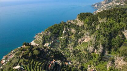 Aerial view of hilly coastal landscape with terraced greenery and scattered buildings along the coastline of a clear blue sea, reminiscent of the stunning Amalfi coast and perfect for summer adventures.