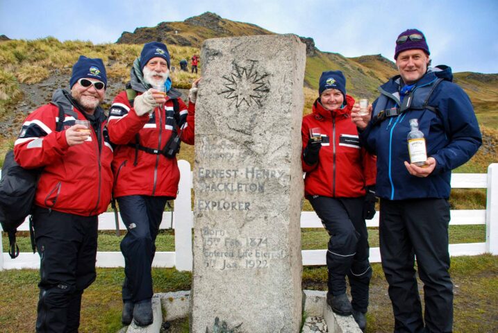 Four people in red jackets and blue hats stand beside a monument memorializing explorer Ernest Henry Shackleton, each holding a drink and smiling, with a hilly Antarctic landscape in the background—a perfect blend of travel and exploration.