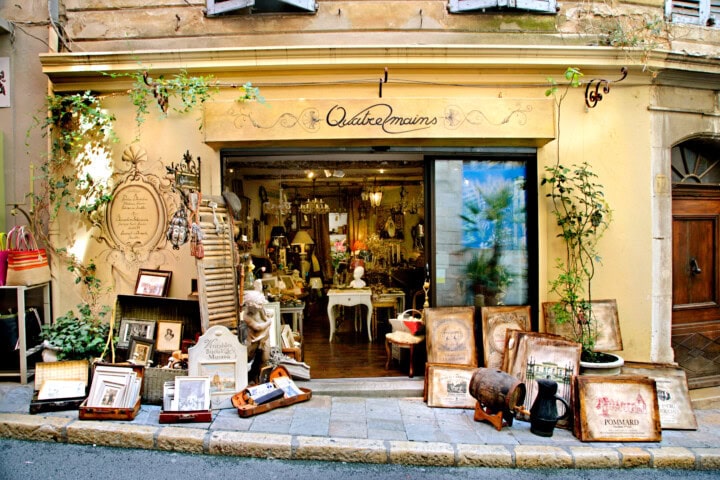 A quaint antique shop in France with a variety of vintage items, paintings, and decorative objects displayed outside its entrance on a cobblestone street. The store's name, "Quatre mains," is shown above the door.