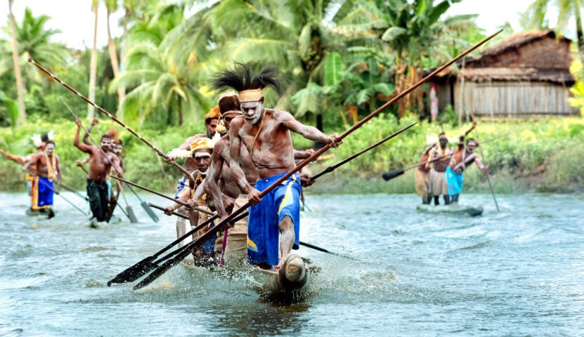 A group of men in traditional attire and face paint paddle canoes with long spears on a river in Papua New Guinea, surrounded by lush tropical vegetation and wooden huts.