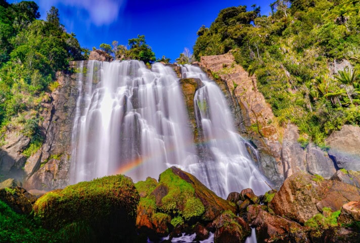 A large waterfall cascades over a rocky cliff surrounded by lush greenery in New Zealand. A faint rainbow forms at the base of the falls under a clear blue sky.