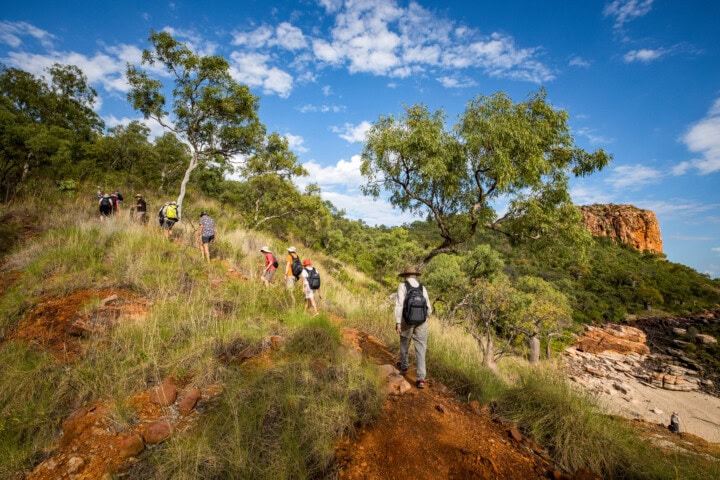 A group of hikers walks up a rocky, grassy hill surrounded by trees, with a large orange rock formation in the background under a partly cloudy Australian sky.