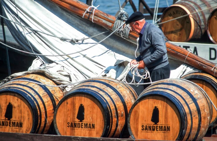 A man in a beret and dark clothing handles rope on a boat near several wooden barrels labeled "Sandeman," evoking the rich maritime heritage of Portugal.