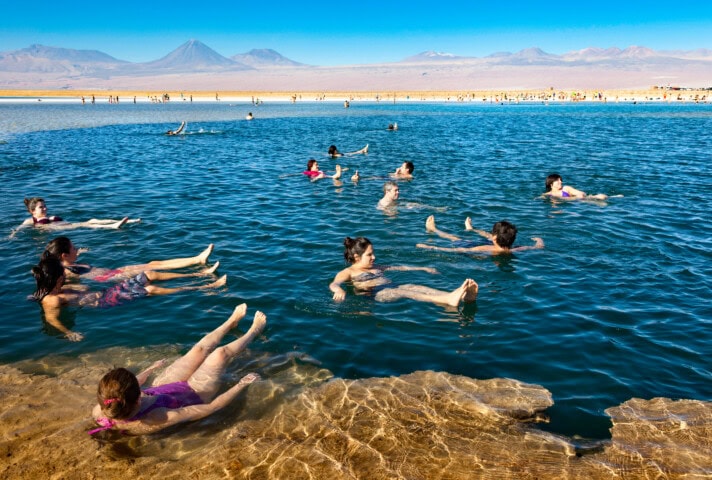 People float and swim in a tranquil, clear blue lake surrounded by a vast, sandy landscape with the mountains of Chile in the background.