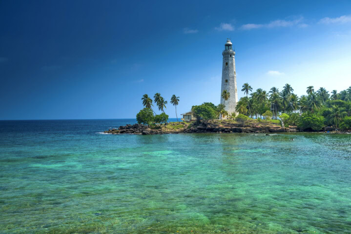 A tall lighthouse stands on a small rocky point surrounded by palm trees, overlooking clear turquoise waters, with a deep blue ocean and sky in the background—a perfect travel spot showcasing the beauty of Sri Lanka's tourism treasures.
