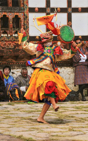 A dancer in a colorful costume and mask performs a traditional Bhutanese dance outdoors, with an audience sitting and standing behind.