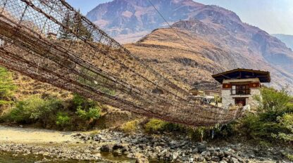 A long suspension footbridge made of rope and wood crosses a rocky river, with a traditional building and dry mountains in the background—a perfect scene for hiking in Bhutan on your cultural journey.