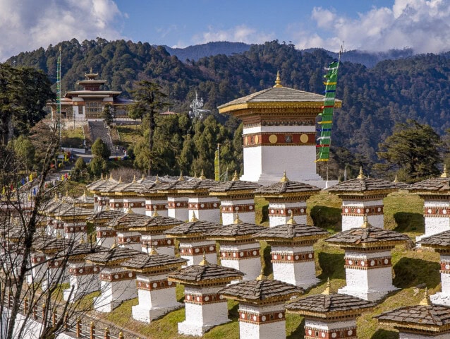 A series of white stupas with golden spires are arranged on a hillside in Bhutan, offering a picturesque sight for tourism. A large temple with traditional architecture is seen in the background amidst a lush green forest and mountains, making it an enchanting travel destination.