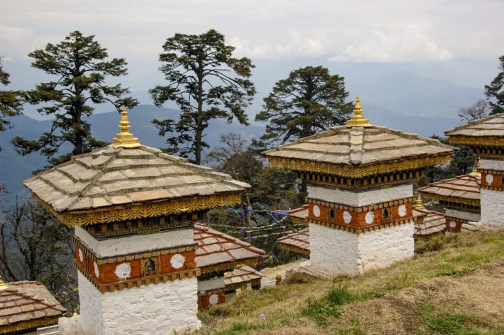 A row of traditional stupas with white walls and golden spires adorns the mountainous landscape of Bhutan, surrounded by tall trees under a cloudy sky—an inviting sight for any travel and tourism enthusiast.