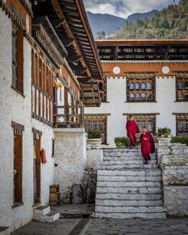 Two monks in red robes stand on the steps of a traditional Bhutanese building with white walls and intricate wooden details, set against a picturesque mountainous backdrop.