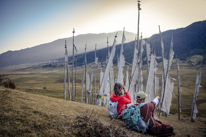 Two people sitting on a hillside in Bhutan, surrounded by tall white prayer flags, with a scenic valley and mountains in the background—a perfect snapshot of travel and tourism.