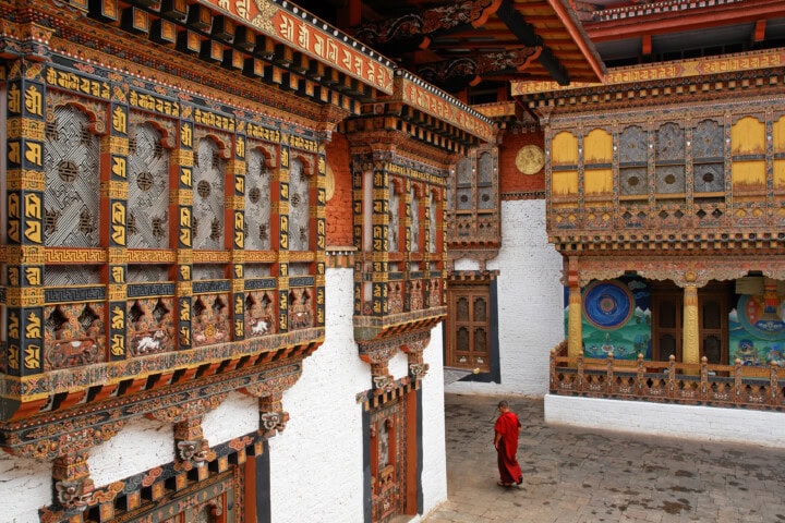 A person in a red robe walks through a Bhutanese courtyard featuring ornately decorated buildings with intricate woodwork and colorful paintings. The traditional architecture showcases detailed designs and patterns.