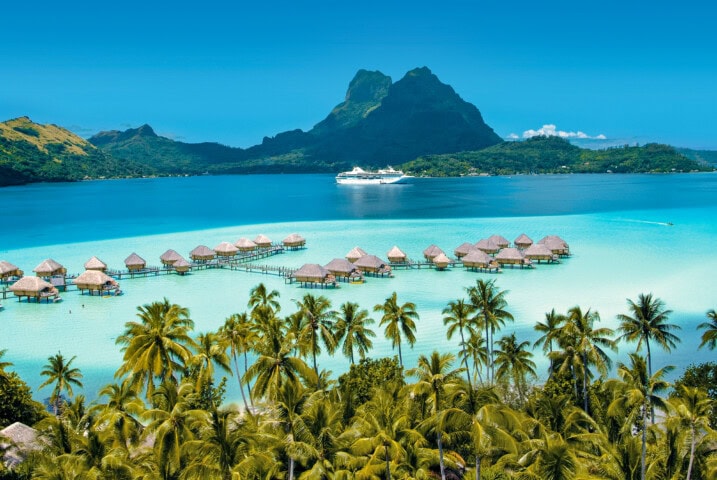 Scenic view of overwater bungalows on a turquoise lagoon in French Polynesia with a cruise ship in the distance, surrounded by lush palm trees and mountains in the background.