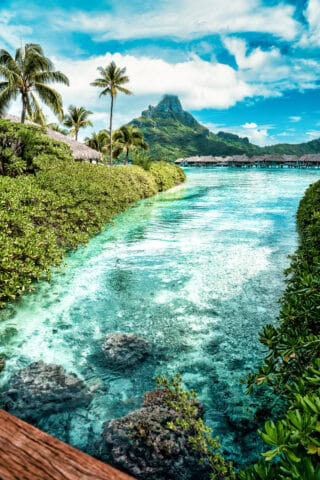 Tropical landscape featuring clear turquoise water, lush green vegetation, palm trees, overwater bungalows, and a mountain under a partly cloudy sky in French Polynesia.