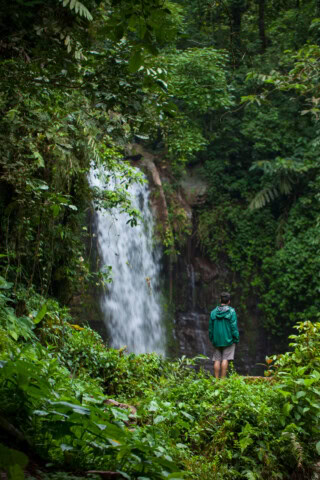 A person in a green jacket stands near a waterfall in a lush, green forest, capturing the beauty of Malaysia's breathtaking landscape, a perfect spot for nature enthusiasts and those passionate about travel and tourism.