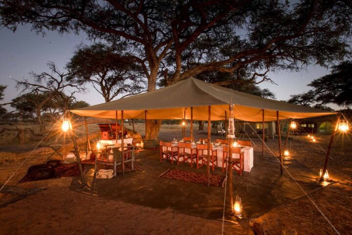 A large canvas tent is set up outdoors with a dining table and chairs underneath it, perfect for an adventure. Lanterns hang around the perimeter, casting a warm glow in the twilight. Trees and a clear sky provide the backdrop for this memorable trip.