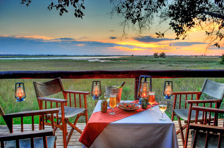 Outdoor dining setup with a table covered by a white cloth and red runner, surrounded by wooden chairs, overlooking a scenic sunset view. Lanterns provide lighting as food and drinks are placed on the table, perfect for travelers to relax after a long hike.