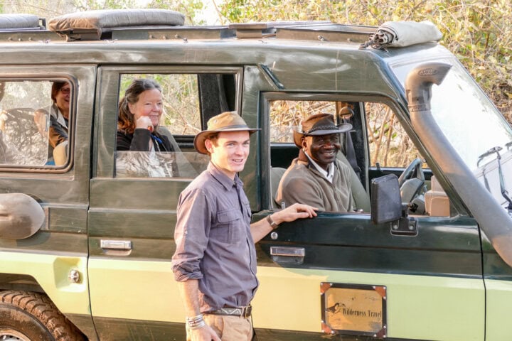 Four people are on a safari in Botswana in an off-road vehicle. Two are sitting in the front seats, one in the back, and one is standing outside next to the open driver's window. They all appear to be smiling, capturing what could be their best adventure for their travel blog.