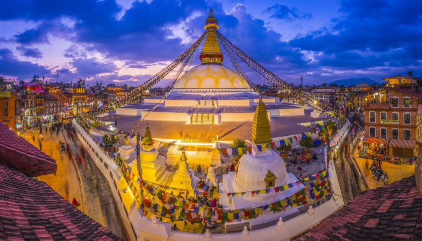 A large stupa decorated with colorful prayer flags in Nepal, surrounded by a bustling square and buildings, under a vibrant evening sky with clouds at twilight.