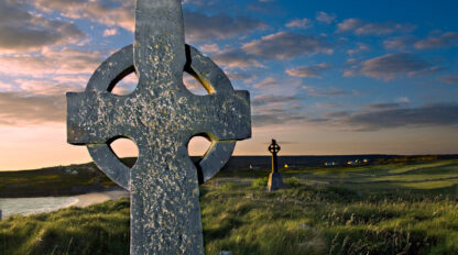 A weathered stone Celtic cross stands in a grassy field during sunset in Inishmore, Ireland, with another cross visible in the distance.