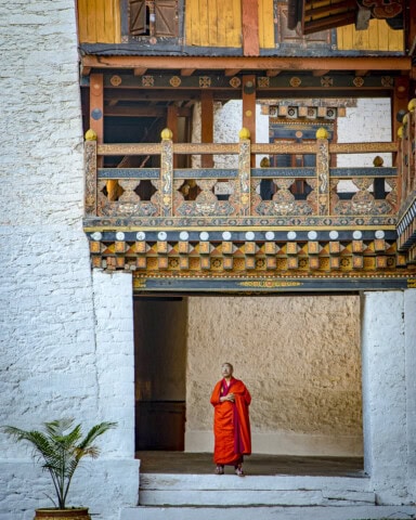 A monk in red robes stands beneath an ornately decorated wooden balcony in an outdoor courtyard, beside a stone wall and a small potted plant, evoking the serene beauty of Bhutan and inviting a sense of travel and tourism to this tranquil scene.