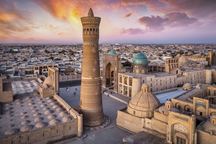 Aerial view of historic city in Uzbekistan with large brick minaret, dome-roofed building, and blue-domed structures at sunset, under a colorful sky.