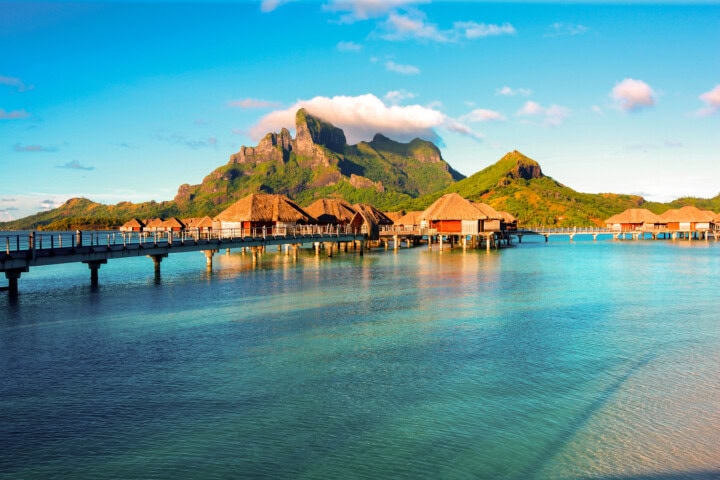 Overwater bungalows on stilts, connected by wooden walkways, sit in a calm turquoise lagoon with a backdrop of lush mountains under a clear blue sky, offering the perfect travel escape to French Polynesia.
