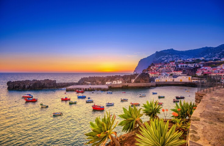 Sunset over a coastal village in Portugal with boats floating in the water, surrounded by rocky cliffs and greenery. The sky transitions from deep blue to warm orange near the horizon.