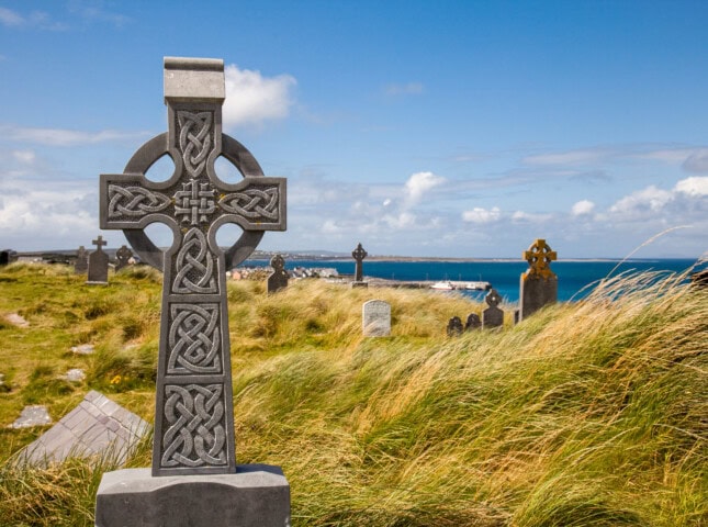 A carved Celtic cross headstone in a grassy cemetery overlooking a coastal landscape in Ireland under a blue sky with scattered clouds.