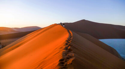 A group of tourists walking along the ridge of a sand dune in Namibia's desert landscape during sunset, with other dunes visible in the background.
