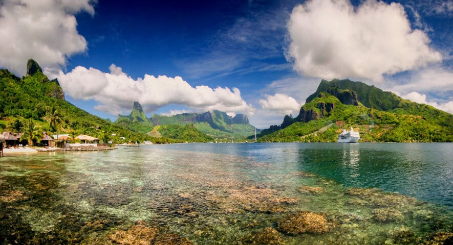 Scenic view of a coastal area in French Polynesia with clear blue water, rocky shoreline, green mountains, scattered buildings, and a large white ship docked near the shore under a partly cloudy sky.