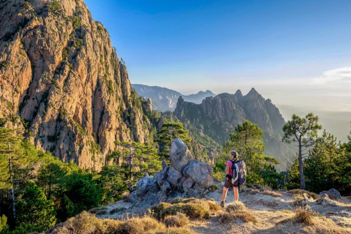 A hiker stands on a rocky ledge in France, overlooking a vast mountain range with steep cliffs and trees under a clear blue sky.