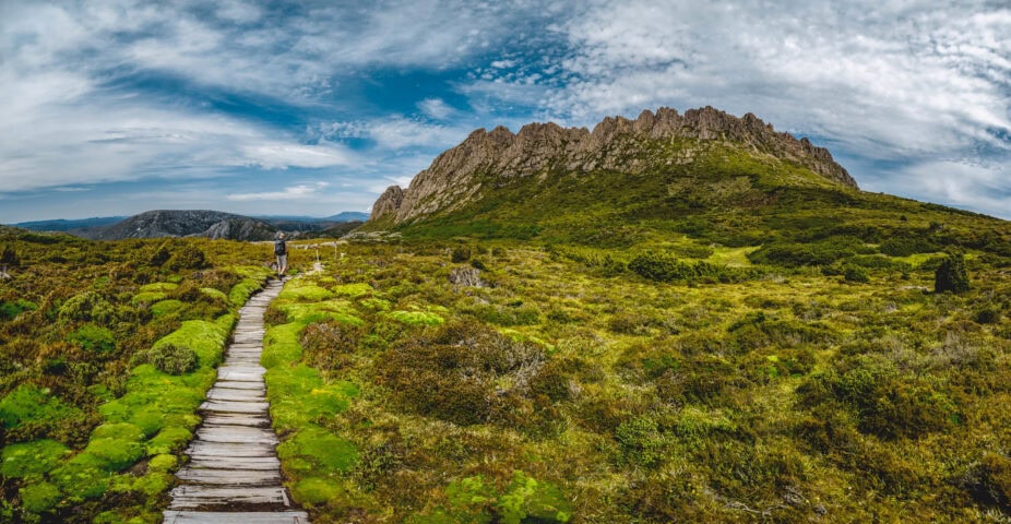 A wooden boardwalk leads through green shrubs toward a rocky mountain under a partly cloudy sky.