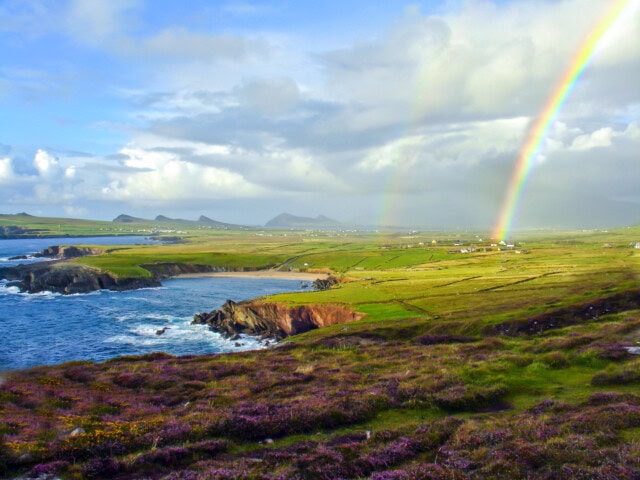 A scenic landscape featuring the coastal beauty of Ireland, with green hills, a rocky shoreline, and a double rainbow stretching into a cloudy sky.