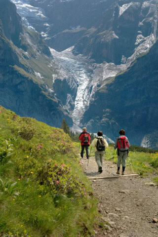 Three hikers wearing backpacks and using trekking poles traverse a trail in the mountainous landscape of Switzerland, with a stunning glacier visible in the background.