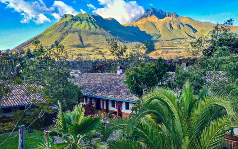 A scenic view of a green valley in Ecuador with a rustic house in the foreground and a majestic mountain range under a partly cloudy sky in the background.