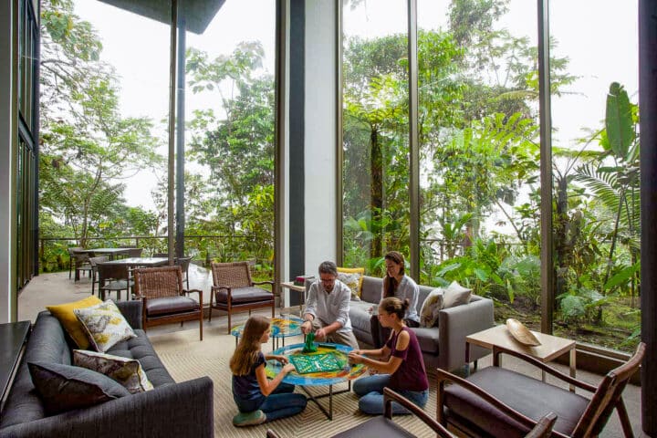 A family of four enjoys a tabletop game in a modern living room with large windows, offering a lush, green view reminiscent of Ecuador's stunning landscapes.