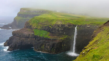 A cliffside waterfall cascades into the ocean under foggy skies in the Faroe Islands. The landscape is green and rugged, with a few buildings visible in the background, perfect for travel reflections.