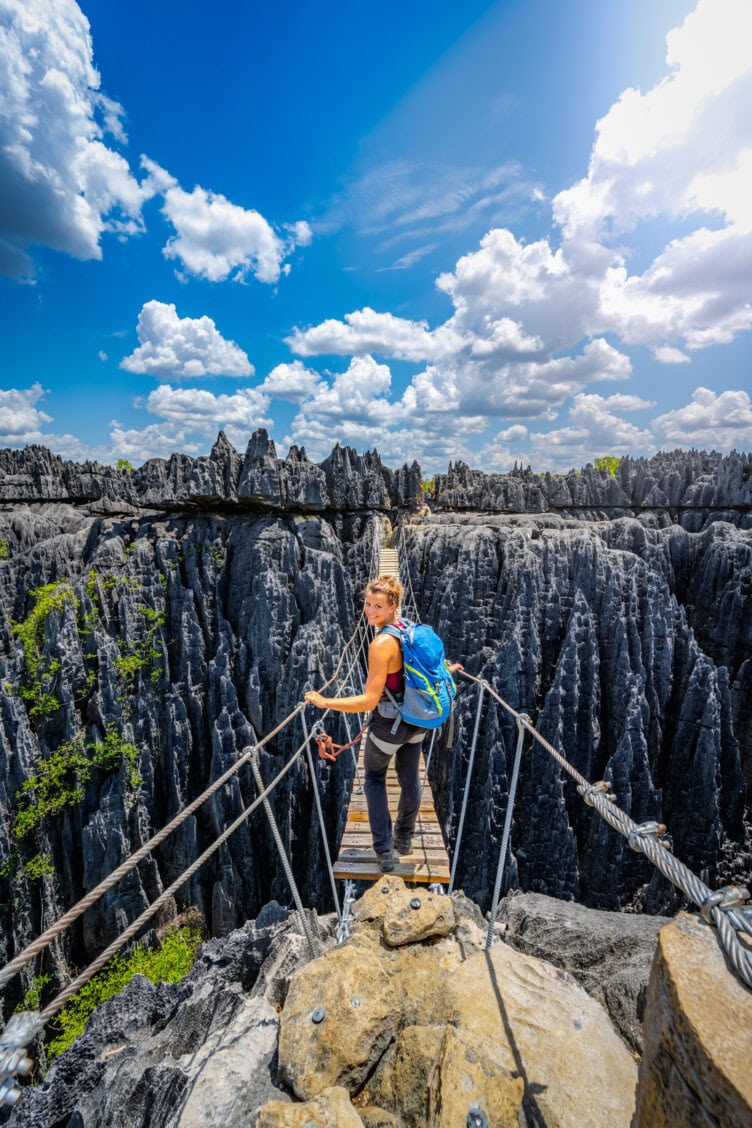 A person with a blue backpack stands on a narrow rope bridge over sharp limestone formations in one of the best parks Madagascar has to offer, beneath a partly cloudy sky.