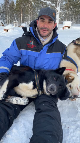 A man in winter gear sits on snow in Finland, holding two resting sled dogs, one black and one white, in his lap.