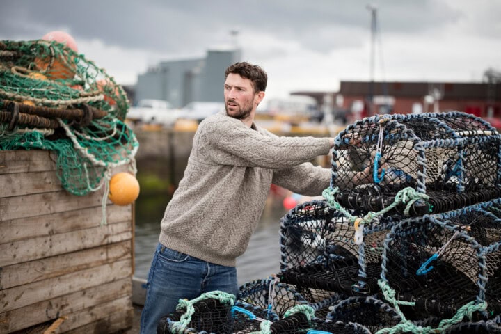 A man wearing a grey sweater and jeans is handling fishing pots near a dock in Scotland, with stacks of fishing nets and equipment in the background. The sky is overcast.