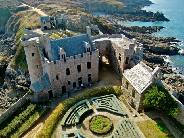Aerial view of a seaside castle in France with a garden featuring a circular hedge centerpiece and patterned pathways, surrounded by rocky cliffs and the ocean—an ideal travel destination for tourism enthusiasts.