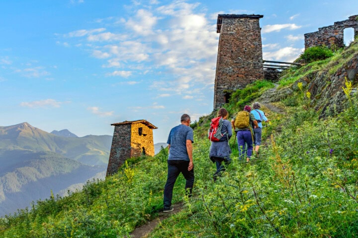 A group of people hike up a grassy path toward rustic stone towers on a hillside in Georgia, with mountains in the background under a partly cloudy sky.