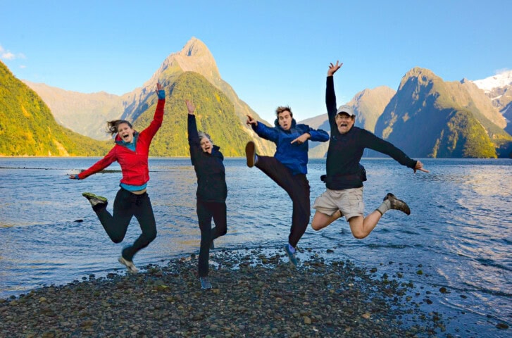 Four people jump in the air on a rocky shoreline with a calm body of water and mountain peaks in the background, capturing the stunning essence of New Zealand.