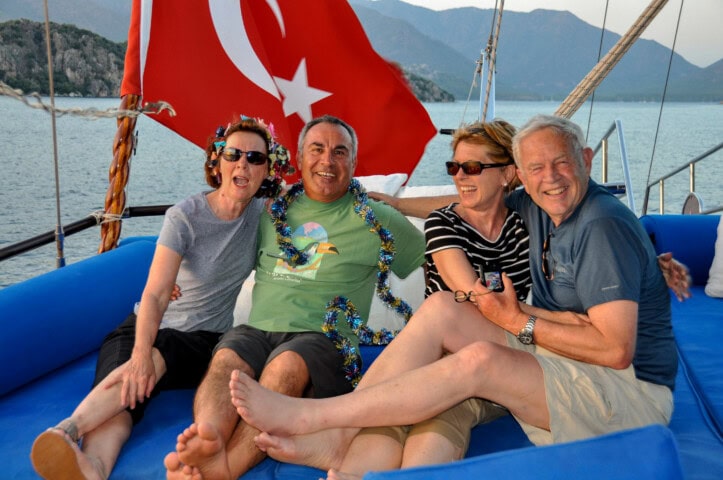 Four people are sitting on a blue boat lounge with mountains and water in the background. They are smiling, with two wearing garlands. A Turkish flag is prominently displayed behind them, capturing the essence of travel and tourism in Turkey.