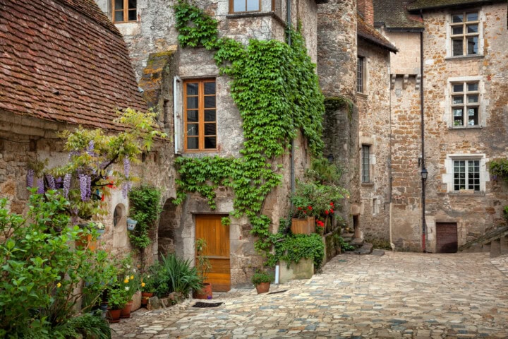 A quaint, narrow cobblestone street in a historic French village with ivy-covered stone buildings and flowering plants in pots along the walls.