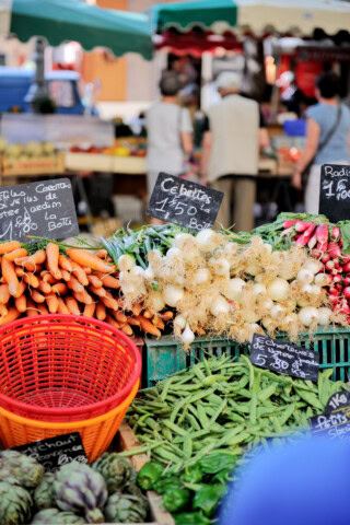 Fresh produce including carrots, onions, radishes, and peas displayed on a market stall in France with a small group of people in the background, inviting travelers to experience authentic local markets and immerse themselves in French culture.